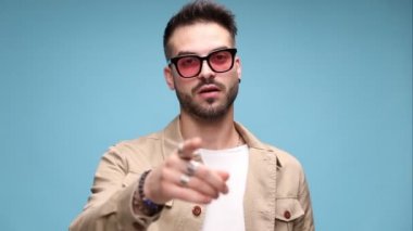 Ceketli, güneş gözlüğü takan seksi genç bir adam. Büyük seksi göğüsler işareti yapıyor ve mavi arka planda flört ediyor.