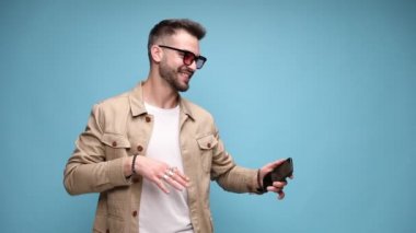 Çekingen, genç, sıradan bir adamın elinde telefon tutup dans etmesi, parmağıyla ekranı göstermesi ve beni işaret etmesi, hareket etmesi ve mavi arka planda dilini dışarı çıkarması.