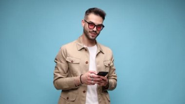 Ceketli, güneş gözlüğü takan mutlu bir adam, yukarı aşağı bakıyor, telefonu tutuyor ve mesaj atıyor, e-postaları okuyor, ekran görüntüsü alıyor, gülümsüyor ve mavi arka planda gülüyor.