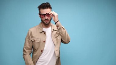 Kendine güvenen, ceketli, güneş gözlüklerini çıkaran, şakakları eğen ve düzelten, mavi arka planda moda pozu veren genç bir manken.