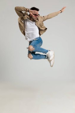 Ceketli, güneş gözlüğü takan, elini havada tutan, yana bakan ve gri arka planda zıplayan seksi genç adam.