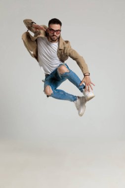 Ceketli, güneş gözlüklü, havalı, genç bir manken. Havaya zıplıyor, ayaklarına dokunuyor, gri arka planda poz veriyor.