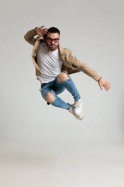 Ceketli, güneş gözlüğü takan, el ele tutuşan ve gri arka planda havaya zıplayan seksi erkek.