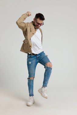 Ceketli, güneş gözlüğü takan, kollarını havaya kaldırmış, zıplıyor ve gri arka planda zaferi kutluyor.