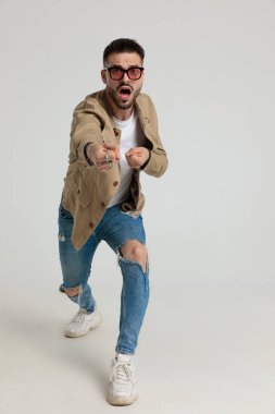 Ceketli, güneş gözlüklü, gergin genç adam ipi çekiyor ve gri arka planda yürüyor.