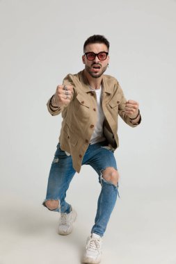 Ceketli kızgın genç adam güneş gözlüğü takıyor, yumruklarını kaldırıyor ve vuruyor, tehdit ediyor ve kavga ediyor, gri arka planda yürüyor. 