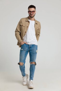 Ceketli seksi genç adam güneş gözlüğü takıyor, el ele tutuşuyor ve gri arka planda yürüyor.