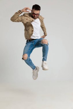 Ceketli, güneş gözlüklü, havalı genç moda adamı, gri arka planda aşağı bakıyor ve havaya zıplıyor.