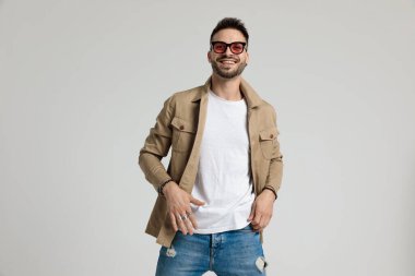 Ceketli, güneş gözlüğü takan, el ele tutuşup gülümseyen, gri arka planda duran mutlu bir adam.