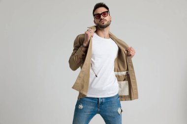 Güneş gözlüğü takan, ceketini tutan ve ayarlayan, gri arka planda moda pozu veren havalı, traşsız genç adam.