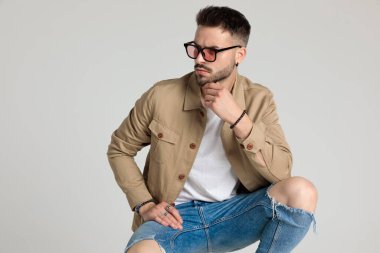 Düşünceli, tıraşsız genç model bej ceket ve güneş gözlüğü takıyor, yan yana bakıyor, çene ele tutuşuyor ve düşünüyor, dirseklerini dizlerinin üstünde tutuyor ve gri arka planda çömeliyor.