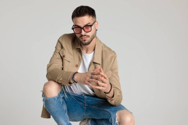 Ceketli genç adam güneş gözlüğü takıyor, parmaklarına dokunuyor, dirseklerini dizlerinin üzerinde tutuyor ve gri arka planda çömeliyor.