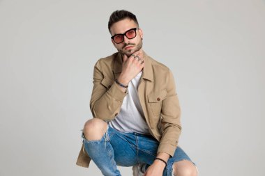 Ceketli, güneş gözlüğü takan, çenesine dokunan ve düşünen, dirseklerini dizlerinin üstünde tutan ve gri arka planda çömelmiş düşünceli genç bir adam.