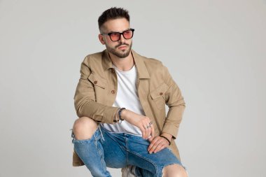 Ceketli, güneş gözlüğü takan, gözlerini kaçıran, dirseklerini dizlerinin üstüne çöküp gri arka planda çömelmiş çekici genç bir adam.