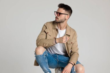 Ceketli seksi genç adam, gözlük takıyor, yan bakıyor, dirseklerini dizlerinin üzerinde tutuyor ve poz veriyor, gri arka planda çömeliyor.