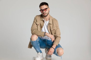 Ceketli, güneş gözlüğü takan, dizlerinin üstünde moda pozu veren, dizlerini büken ve gri arka planda çömelmiş genç manken.
