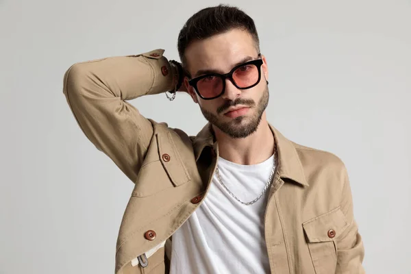 sexy young casual man in jacket wearing sunglasses, holding hand behind neck in a fashion pose and posing on grey background