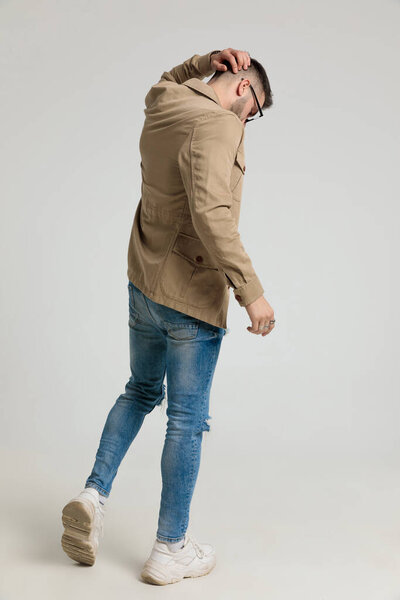 young casual guy in jacket looking down, walking and scratching head on grey background