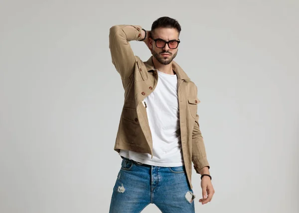 cool fashion model in jacket wearing sunglasses holding hand behind neck and posing, standing on grey background