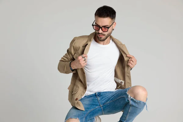 young casual unshaved model wearing sunglasses, looking down and crouching, adjusting jacket and posing on grey background