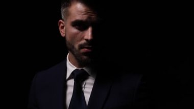 Takım elbiseli genç işadamı gölgeli bir ışıkta duruyor ve poz veriyor, el ele tutuşuyor ve dudaklarına dokunuyor, sessiz bir jest yapıyor, yan yana hareket ediyor, poz veriyor ve davet ediyor, benimle geliyor, yürüyor ve karanlıkta kayboluyor.