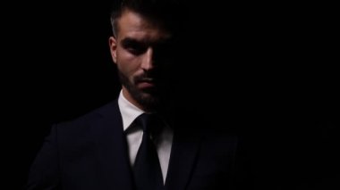 Takım elbiseli çekici genç manken, gölgeli bir ışıkta duruyor ve kravatı düzeltiyor, koyu arkaplanda kollarını kavuşturup poz veriyor.