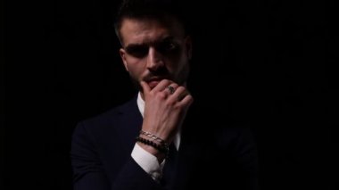Takım elbiseli genç işadamı dudaklara dokunuyor ve hayranlık duyuyor, kafasını oynatıyor ve karanlık arka planda analiz ediyor.
