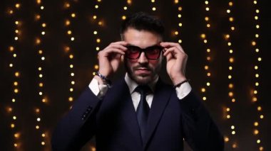 Şık takım elbiseli genç bir iş adamı güneş gözlüğü takıyor ve arka planda poz veriyor.