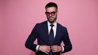 Genç iş adamı güneş gözlüğü takıyor, aşağı bakıyor ellerini ceketinin düğmesine basıyor, kravatını düzeltiyor, parmaklarını saçlarını tarıyor, güneş gözlüğünü takıyor, parmaklarını şıklatıyor, pembe arka planda arıyor ve gidiyor.