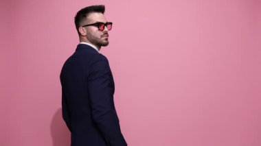 Güneş gözlüğü takmış, bir elini cebine sokan, pembe arka planda sakalını ovuşturan bir iş adamının arka planı.