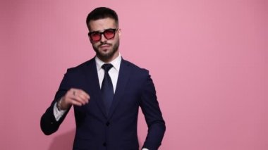 Çekici iş adamı ceketini düzeltiyor, aşağı bakıyor ve gözlüklerini ayarlıyor, kollarını kavuşturuyor, iki parmağını, bir parmağını ve sıfır parmağını pembe arka planda gösteriyor.