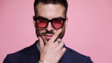 Güneş gözlüğü takan, sakalına dokunan, çenesini kapayan, arkasını dönüp pembe arka planda ayrılan havalı seksi bir iş adamının fotoğrafını çek.