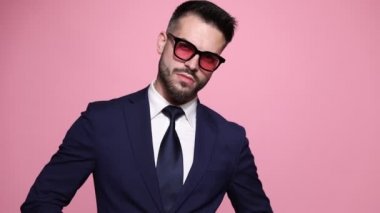 sert moda işadamı kravatını düzeltiyor, güneş gözlüğü takıyor, şık poz veriyor, yanakları şişiriyor, dili dışarı çıkıyor ve pembe arka planda takılıyor.