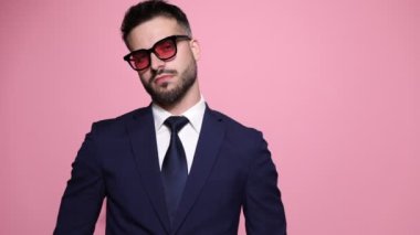 havalı moda işadamı pembe arka planda poz veriyor, güneş gözlüğü takıyor, kenara çekiliyor ve ceketini düzenliyor, 