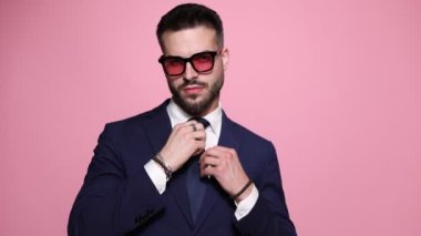 Zarif moda işadamı kravatını ve ceketini düzeltiyor, iki eliyle gülümsüyor ve ileri doğru işaret ediyor, sonra parmaklarını şaklatıyor, yana dönüyor ve pembe arka planda ayrılıyor.