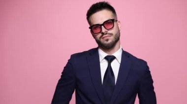 Genç yakışıklı işadamı kameraya bakıyor ve şık bir şekilde poz veriyor, bileziğini takıyor, güneş gözlüklerini çıkarıyor, onlara bakıyor, sonra tekrar takıyor, davet ediyor, pembe arka plana sırtını dönüyor.