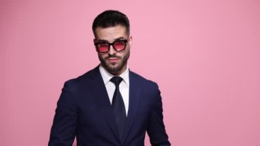 Tavırları olan iş adamı güneş gözlüğü takıyor, kameraya bakıyor, poz veriyor, baştan çıkarıcı bir şekilde dokunuyor ve dudaklarını ısırıyor, pembe arka planda kameraya sırtını dönüyor.