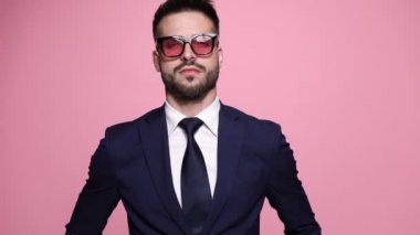 Havalı güneş gözlüğü takan seksi ve rahat iş adamı ceketini düzeltiyor, tavırlarıyla poz veriyor, gözlüğünü ayarlıyor, parmağını dudaklarına dayayıp işaret ediyor, sonra kenara çekiliyor ve pembe arka planda yolunu değiştiriyor.