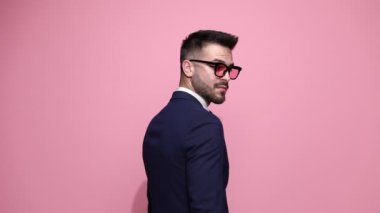Yakışıklı moda işadamı kameraya arkasını dönmüş ve yana bakıyor, yavaşça vücudunu yana çeviriyor, ellerini bir arada tutuyor, ceketini düzeltiyor ve pembe arka planda yüzünde bir gülümsemeyle poz veriyor.