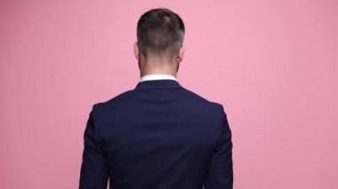 Donanma takım elbiseli genç adam kameraya arkası dönük duruyor, yavaşça başını yana çeviriyor, güneş gözlüğü takıyor ve pembe arka planda havalı bir tavrı var.