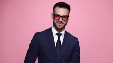 Davranışları olan bir iş adamı ceketini düzeltir, kravatını düzeltir ve başka yere bakar, parmaklarını saçlarını tarar, güneş gözlüğünü kaldırır, kenara çekilir, birini arar ve pembe arka planda bırakır.