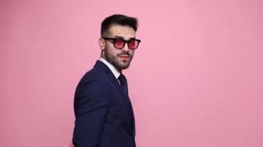 Güneş gözlüklü genç yakışıklı iş adamı arkasını kameraya dönük duruyor, ellerini kavuşturuyor, parmaklarını uzatıyor pembe arka planda kibirle sakalını gezdiriyor.