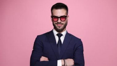 Genç sert iş adamı kollarını kavuşturuyor, güneş gözlüğü takıyor, gülümsüyor, ceketini düzeltiyor, tavırlarıyla poz veriyor, güneş gözlüklerini ayarlıyor, sonra da pembe arka planda arkasını dönüyor.