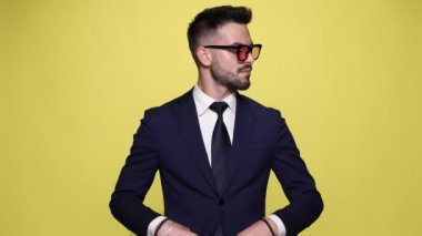Takım elbiseli zarif genç işadamı güneş gözlüğü takıyor, takım elbise bakıyor, kollarını kavuşturuyor, gözlük takıyor, gülümsüyor, parmakla işaret ediyor, ceketini ayarlıyor ve sarı arka planda yürüyüp gidiyor.