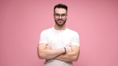 Beyaz tişörtlü ve gözlüklü yakışıklı bir adam, kolları pembe arka planda çapraz duran, sonra kameraya bakan, gülümseyen ve pembe arka planda yumruklarını kaldıran.