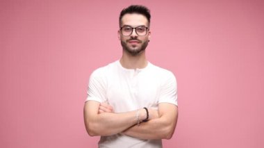 Kollarını kavuşturmuş, gözlük takmış, başını kaldırıp gülümseyen, pembe arka planda dans eden elleri olan genç ve yakışıklı bir adam.