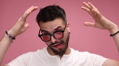 Güneş gözlüğü takan, sakala dokunan, aptal gibi ellerini havada sallayan, dilini dışarı çıkaran, gözlüğünü kaldıran ve pembe arka planda bir eliyle kafasına el sallayan genç bir adamın yakınına.