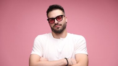 Kolları çapraz pembe arka planda duran, güneş gözlüğü takan, omuzlarını silkeleyen ve birinin neden bahsettiği hakkında en ufak bir fikri olmadığını söyleyen genç bir adam.