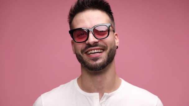 ハンサムなカジュアルな男が笑顔でサングラスをかけ顔を真剣にし髭に触れ目をそらしピンクの背景を覗かせ — ストック動画