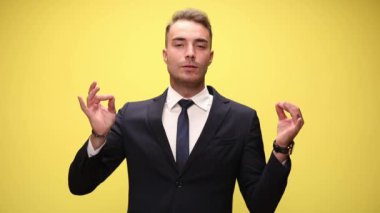 Parlak takım elbiseli genç işadamı, gülümsüyor, parmaklarını mudra pozisyonunda tutuyor, kollarını ve vücudunu rahatlatıyor ve sarı arka plan üzerinde meditasyon yapıyor.